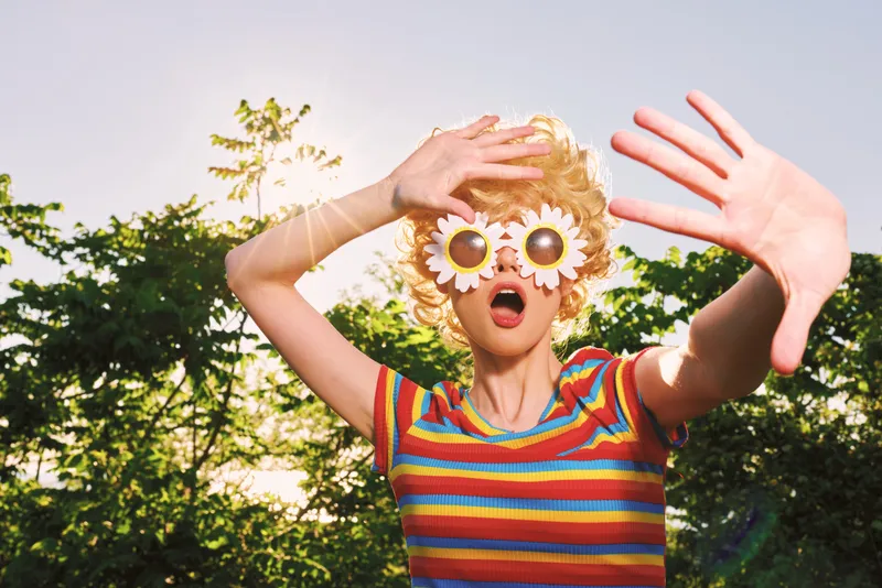 A playful woman in a striped shirt outdoors wears whimsical flower sunglasses, posing with arms raised. Bright retro colors evoke carefree summer vibes.