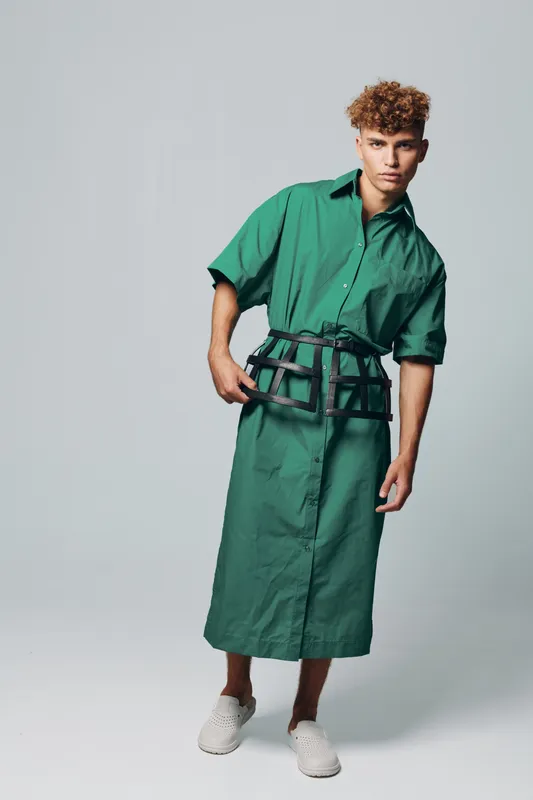Young man with curly hair wearing long green shirt dress and black belt posing in studio with neutral background. Fashion model standing with casual white shoes and serious expression.