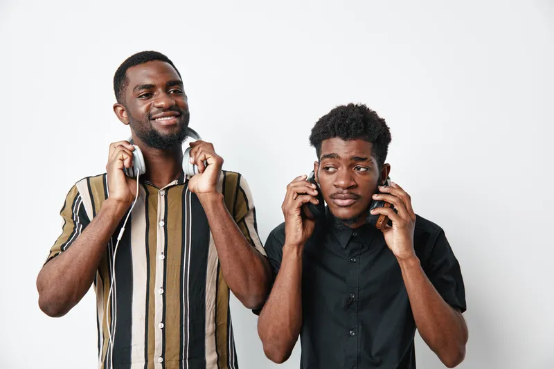 Two young black men with dark skin using smartphones and headphones, standing against white background. Casual clothing and modern technology for communication and entertainment.