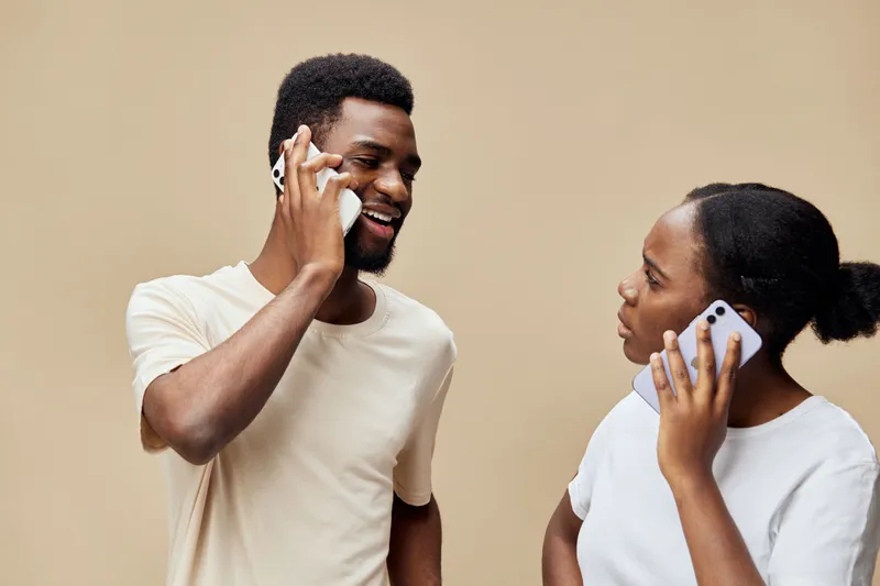 Happy African couple talking on the phone, wearing casual clothes, set against a neutral beige background, promoting communication and connection