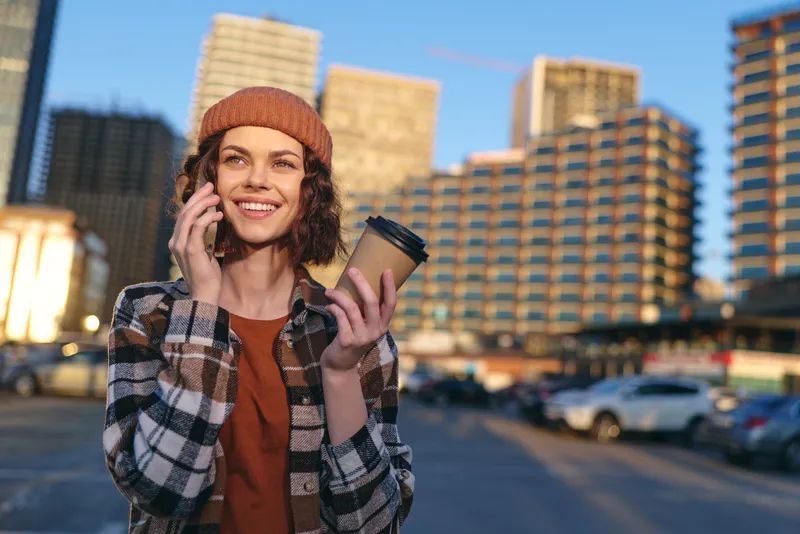 woman with coffee and phone in urban street, smile and candid beanie portrait at golden hour, authenticity and candid lifestyle with golden hour glow, mindful living and emotional storytelling