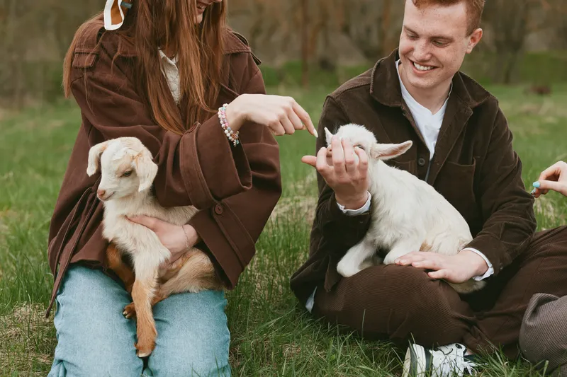Young man and woman sitting on grass holding and playing with two baby goats in a casual outdoor setting during a relaxed lifestyle moment.