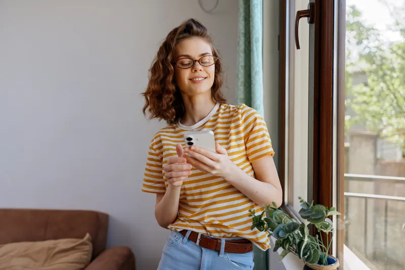 Casual woman at home by a window uses a smartphone, wearing striped shirt and jeans, relaxed lifestyle scene with natural light and cozy interior, she stands beside a potted plant, smiling softly.