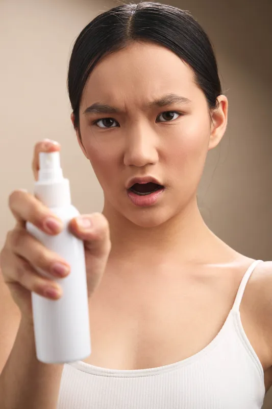 Asian woman in a white tank top stands in a simple setting, holding a spray bottle toward the camera with a surprised expression, wide eyes and open mouth, conveying urgency and immediacy.