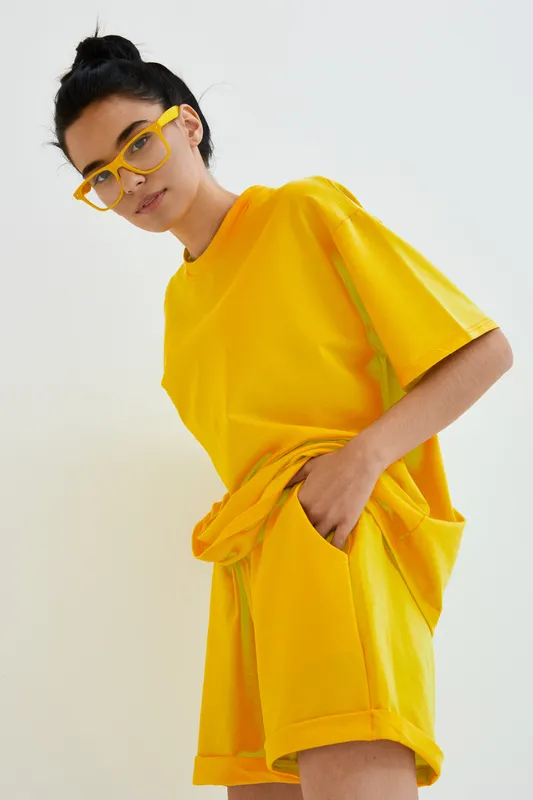 young woman wearing bright yellow casual outfit and matching glasses, posing with hands in pockets against plain light backdrop, modern street style portrait