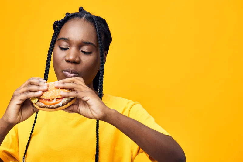 Young Black woman enjoying a tasty burger against a bright yellow background, wearing a casual yellow T shirt Emotion Delight in fast food