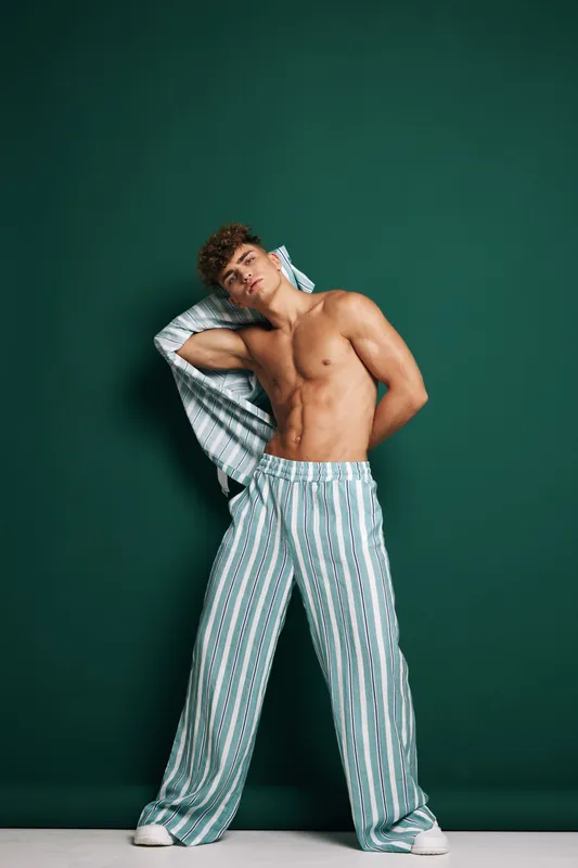 Young man posing shirtless in striped pajama pants with open shirt against dark green background. Casual fashion, relaxed style and confident male model in studio setting.