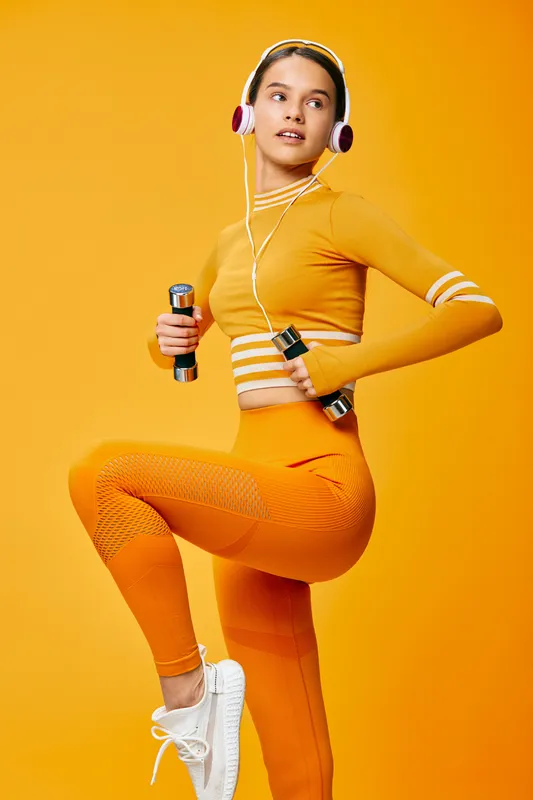 Female fitness model in bright orange activewear holding small dumbbells and wearing headphones, striking a dynamic studio workout pose against a vibrant orange background, energetic and focused.