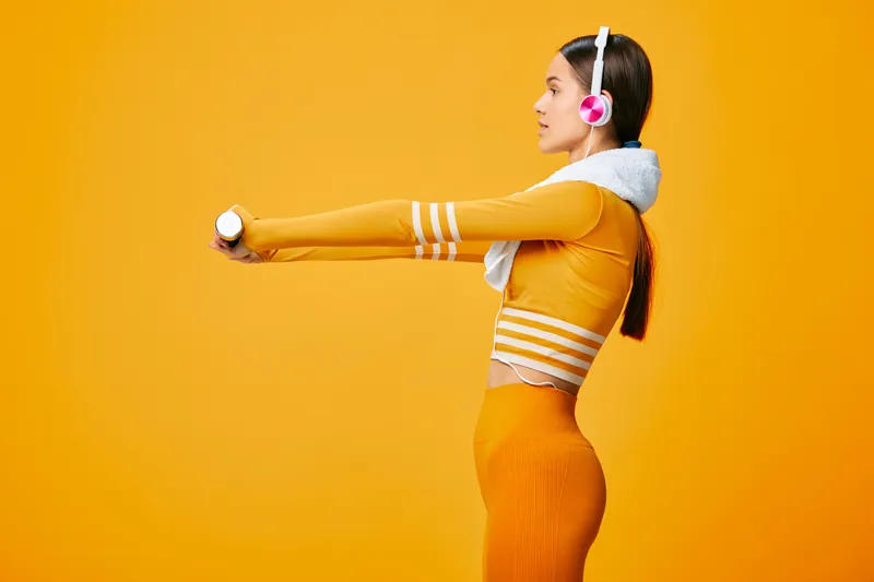 Woman fitness athlete stretches arms in a dynamic pose, wearing orange athletic wear and headphones against a bright background, exuding energy, focus and healthy lifestyle.
