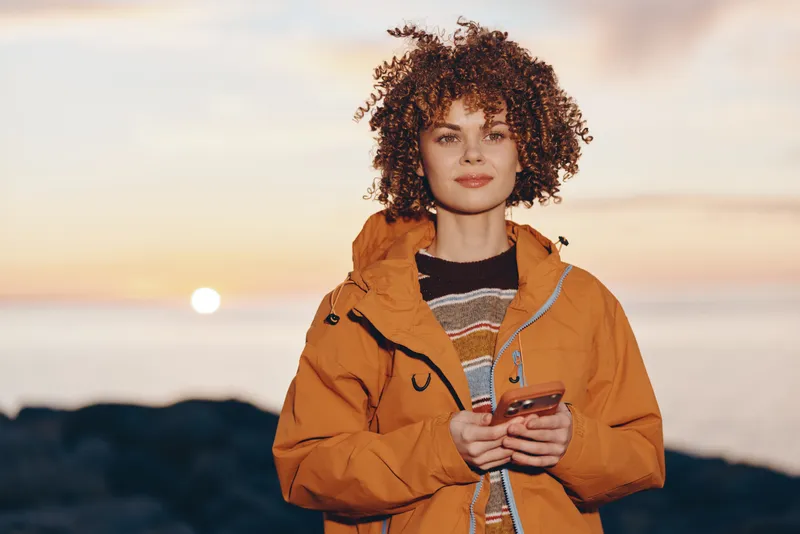 Lifestyle woman in a rust orange jacket stands outdoors at sunset by the sea, holding a coffee mug and smiling softly, curly hair frames her face, warm evening light in the breeze