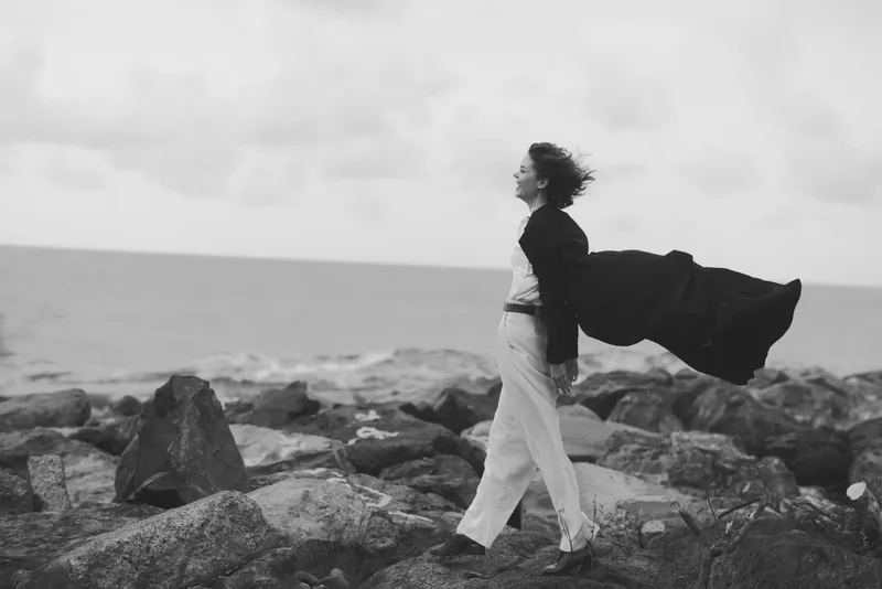 Wind swept coat flows as a woman walks along jagged shore by the sea, capturing motion, solitude, and freedom in a moody black and white landscape