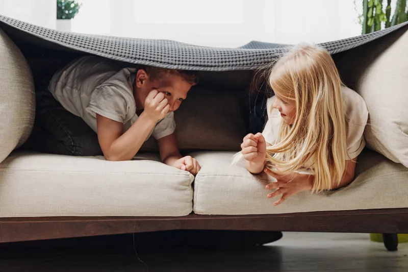 Two children play hiding under the sofa cushions in a cozy living room, smiling and enjoying a playful moment together.