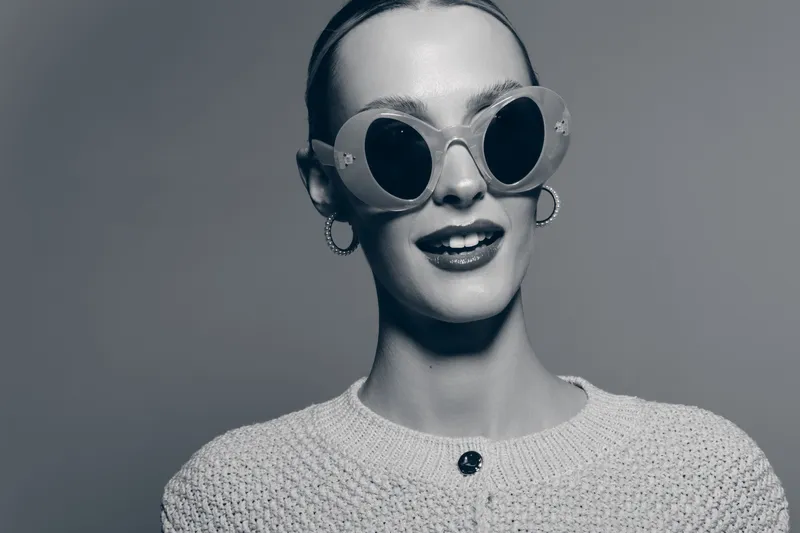 Young white woman with pale skin wearing round sunglasses and hoop earrings smiles confidently. Studio portrait with neutral background and trendy fashion style.