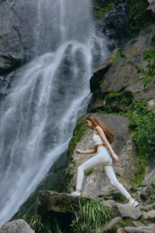 Active woman hiking near a majestic waterfall in white sportswear, surrounded by lush green vegetation and rocky cliffs, embodying outdoor adventure and fitness.