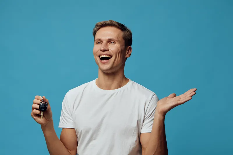 Joyful smiling young man with short brown hair wearing casual white t-shirt holding car key keychain and gesturing with hand while standing against bright blue background studio portrait. People lifes