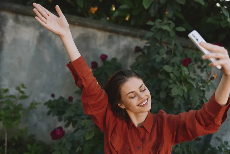 Smiling woman in a red shirt poses outdoors, arms raised in joy. Close-up selfie vibe with garden backdrop, capturing a vibrant lifestyle and carefree mood