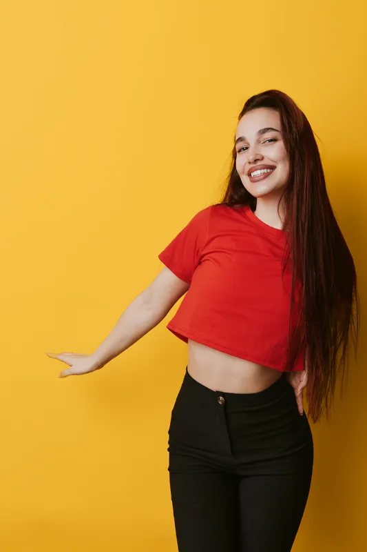 Young woman with long hair wearing red crop top and black pants posing smiling against yellow background. Casual style, happy expression and confident posture for lifestyle portrait.