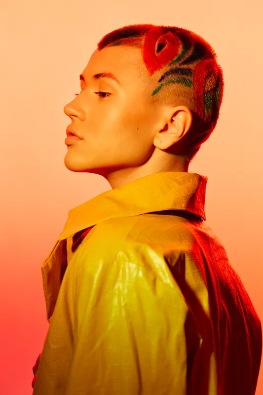 young woman with artistic shaved hairstyle featuring red roses, wearing yellow jacket against orange gradient background in studio lighting
