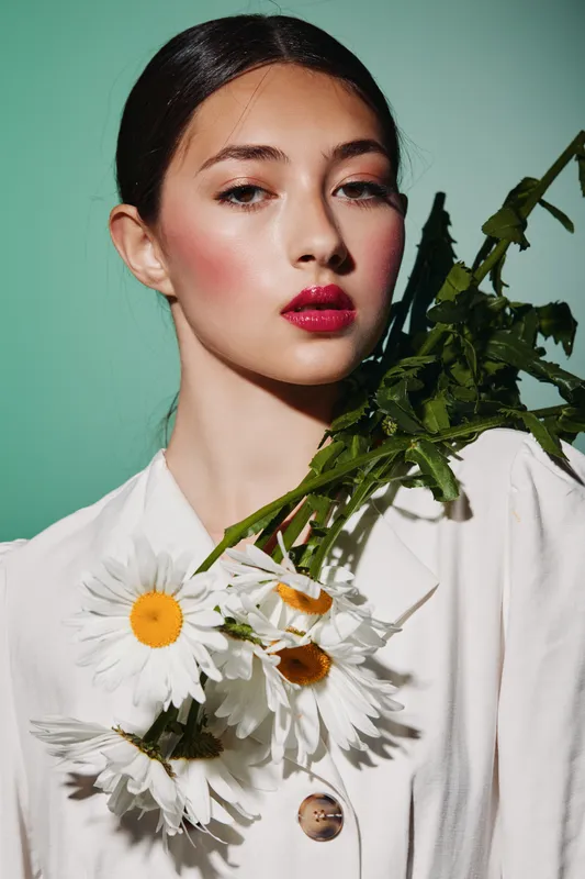 Fashion model with bright red lipstick poses with white flowers over shoulder against green background. Creative beauty concept with vivid makeup and styling.