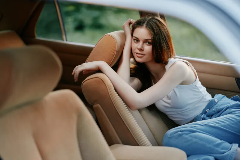 Young woman in casual outfit relaxing in a car, smiling softly, with natural green background and warm tones, conveying a sense of enjoyment and freedom.