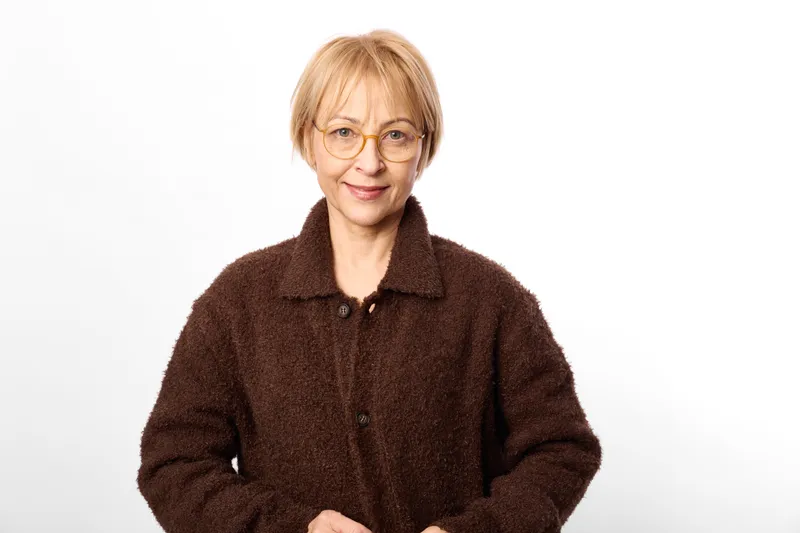 Woman mature senior portrait in studio, glasses and brown sweater, standing calm against white background, gentle expression, soft light, minimal composition for aging wellness