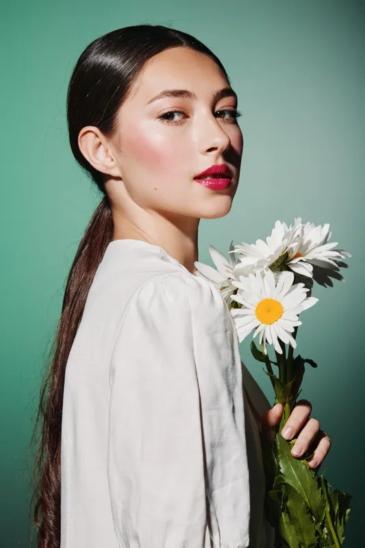 Young woman with glowing skin and bold red lips holding white daisies against a green background. She wears a white blouse and sleek hair drawing beauty, fashion themes.