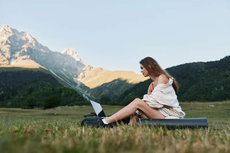 A young woman sits on a yoga mat in a meadow with a laptop, enjoying fresh air and a scenic mountain landscape, creating a calm and productive outdoor work scene.