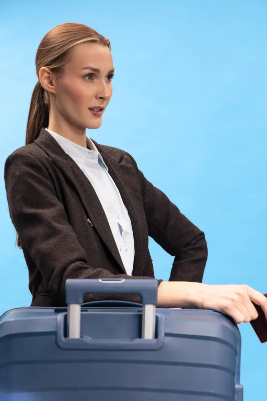 Young woman in formal jacket stands with suitcase and passport against blue background. Concept for travel, business trip, and preparation for flight.