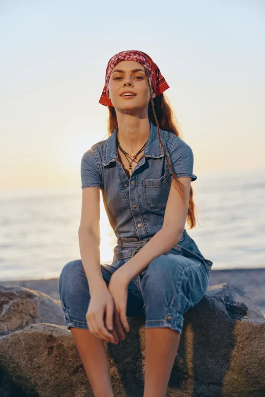 lifestyle woman in modern boho-western denim street style sitting on rocks by the sea with film color tones and relaxed summer vibe