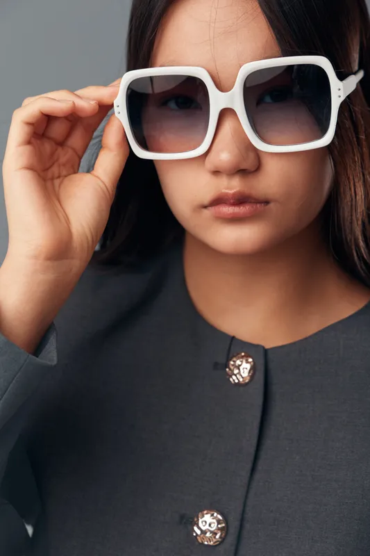 Woman with serious expression and large white sunglasses touching frame, wearing dark coat with decorative buttons against black background in studio closeup portrait.