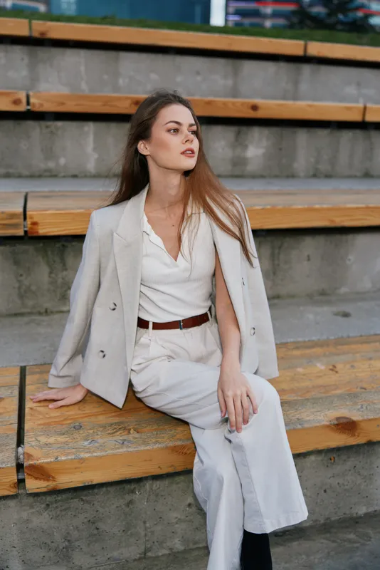 Stylish woman in light business outfit sitting on wooden stairs with confident calm pose and neutral outdoor background in spring or autumn fashion