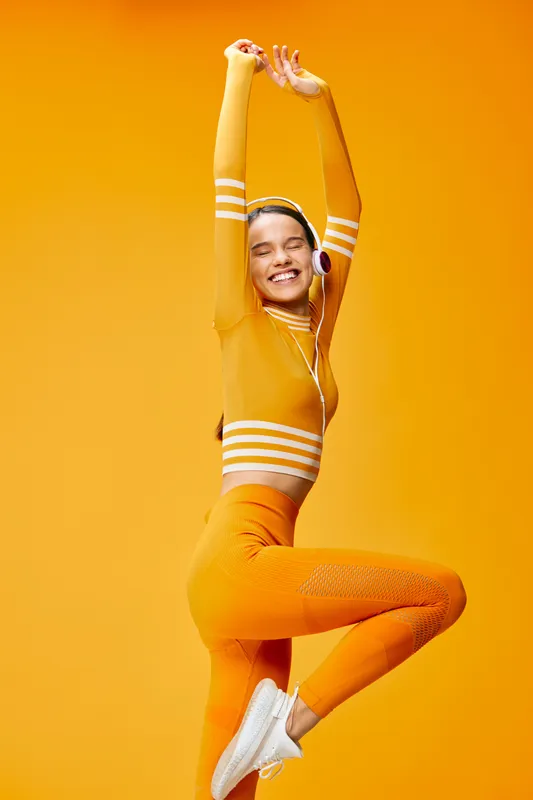 Vibrant portrait of a smiling woman in orange athletic wear mid jump, showcasing fitness energy and sport style with headphones against a bold orange background.
