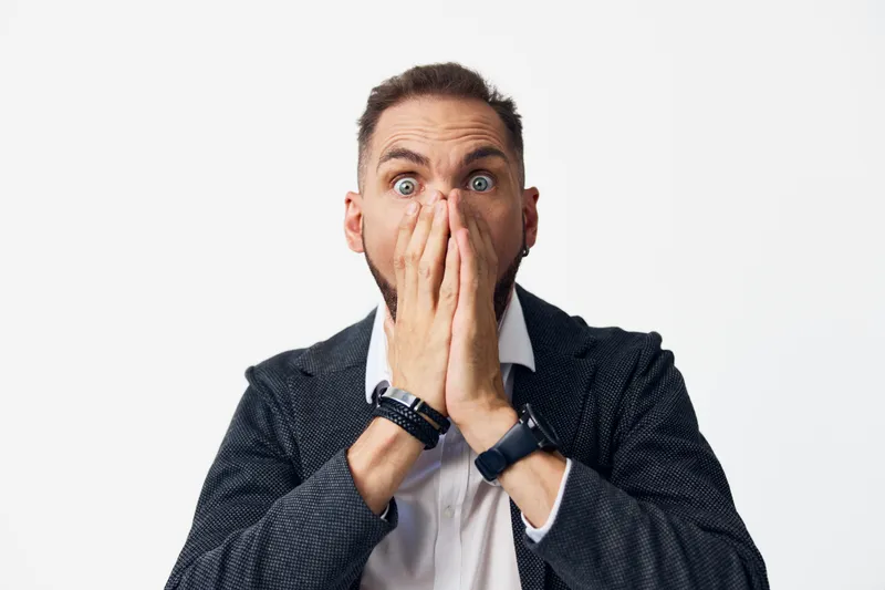 A business professional man shows surprise with hands over his mouth against an isolated white background, conveying unexpected news and authentic emotion.
