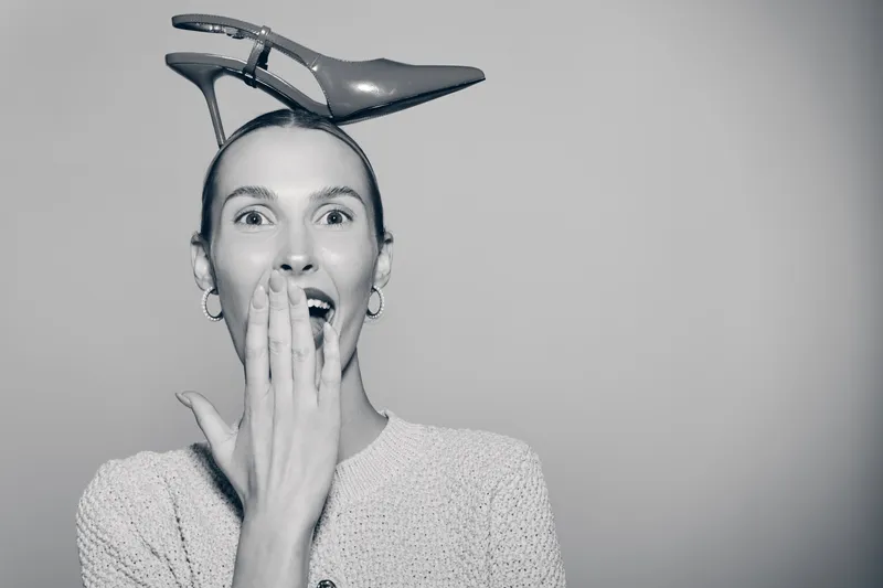 Surprised white woman with light skin and brown hair wears high heel shoe on her head and covers mouth with hand. Concept of humor, fashion, and playful expression.