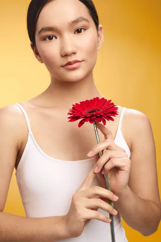 asian woman holds a red flower and gazes softly at the camera against a warm yellow backdrop, conveying calm elegance and a subtle, natural mood in a stylish studio setting.