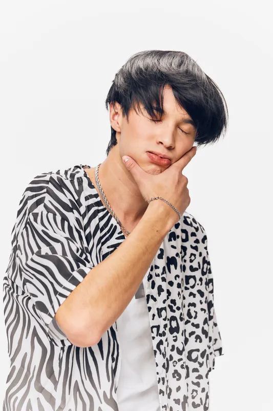 Young Asian man with closed eyes and thoughtful expression wearing a black and white animal print shirt and silver jewelry on a white background.