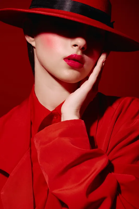 Fashion model in a red outfit with bold red lipstick partly hiding her eyes with a stylish wide-brimmed hat. Creative beauty portrait with dramatic lighting and makeup. Stylish concept.