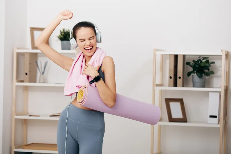 Fitness woman in a bright home studio, wearing headphones, carrying a yoga mat and towel, mid workout with energetic movement and positive lifestyle, showcasing exercise and motivation.