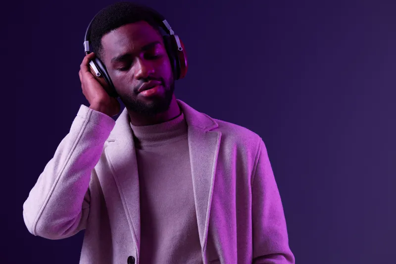 Stylish young African man enjoying music with headphones in a modern urban setting, showcasing relaxation and creativity with a purple background