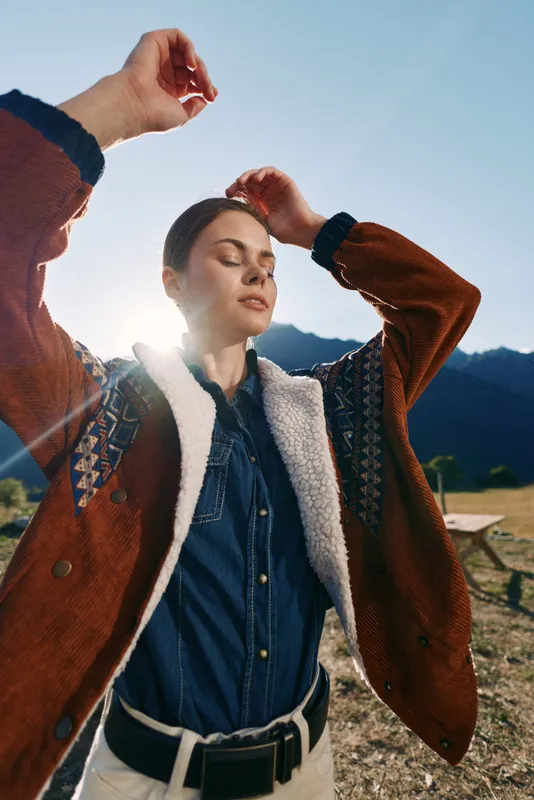 woman outdoors mountains sunlight jacket portrait relaxed denim, young adult in shearling coat with eyes closed, enjoying serene nature, backlight sunflare and scenic landscape during countryside hike