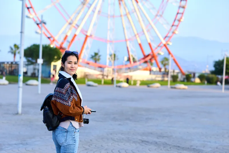 Woman with camera at an outdoor park, standing near a towering ferris wheel, wearing a casual jacket and backpack, capturing travel moments under daylight in a vibrant amusement setting
