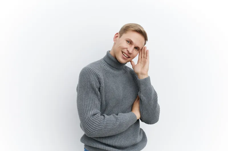 Smiling young caucasian man wear gray knitted sweater hold hand near ear listen carefully isolated on white background concept mental health and gadgets
