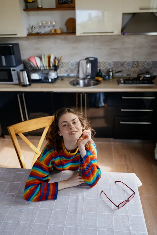 A woman in a colorful striped sweater sits at a table in a bright kitchen, head resting on her hand, glasses on the table. A calm, candid lifestyle moment at home.