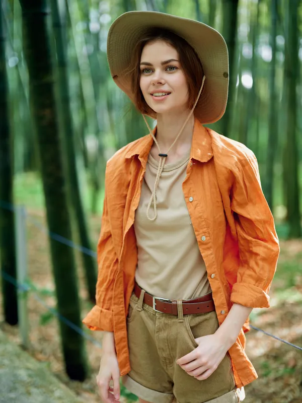Woman in hat and orange shirt walking in bamboo forest, hiking with a smile. Casual travel portrait, nature exploration, outdoor adventure and eco friendly trip vibe.