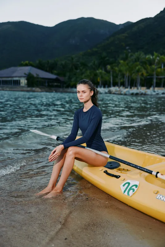 Young woman sitting on a yellow kayak by the shore, wearing dark swimwear, calm expression, tropical landscape with mountains and palm trees in the background.