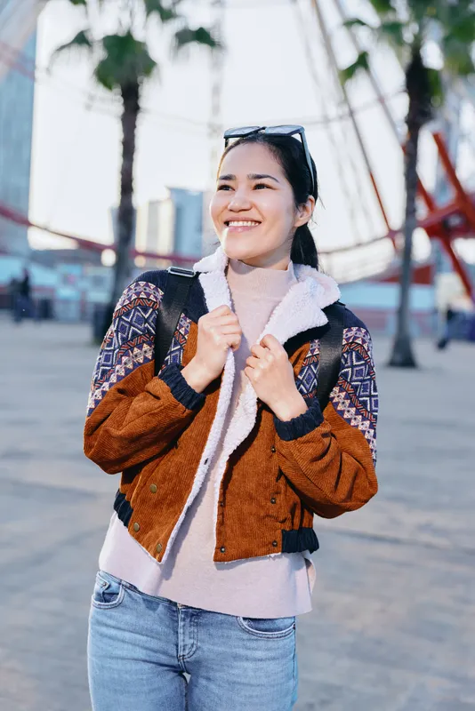 Smiling woman in a warm brown and cream fleece jacket stands on an urban street, casual fashion pose under soft daylight with a modern city backdrop