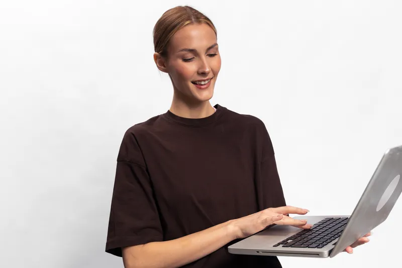 Smiling young woman in dark shirt holding laptop and typing, isolated on white background. Concept of remote work, technology use, and modern communication.