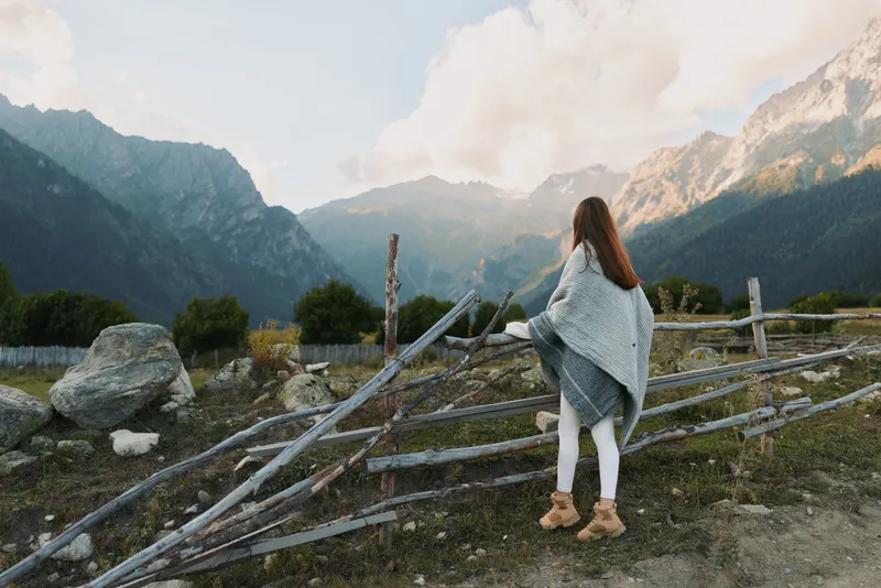 Woman lifestyle traveler wrapped in a blanket standing near wooden fence with mountain view. Outdoor autumn scene with cloudy sky and natural landscape background.