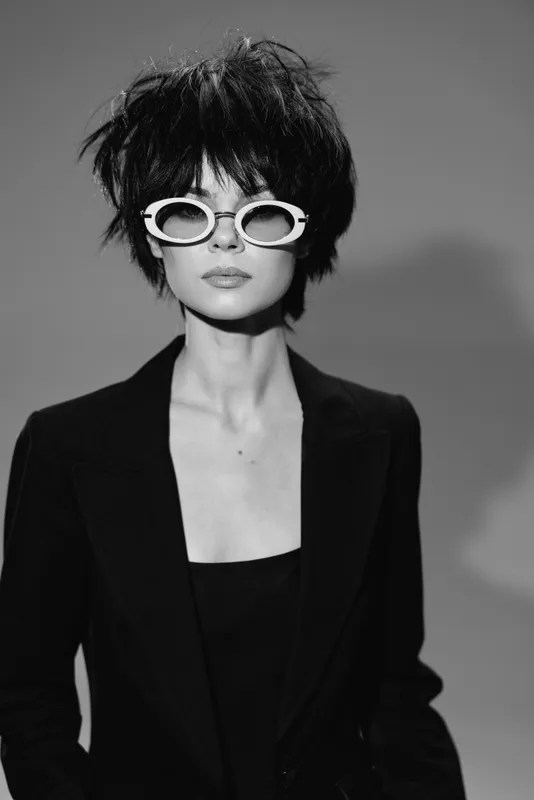 Black and white portrait of a stylish woman wearing white sunglasses and a black outfit with short messy hair.