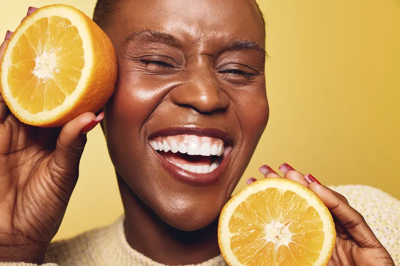 Close-up of smiling black woman with dark skin holding two orange halves near face on yellow background. Concept of healthy lifestyle, vitamin C, skincare, and natural beauty.